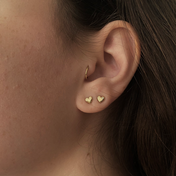 a woman wearing gold stud heart earrings, one of them is set with a diamond